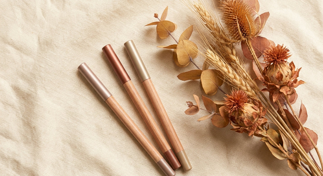 A styled flatlay of eyebrow pencils in warm auburn and taupe shades arranged on cream linen with dried copper-toned botanicals.
