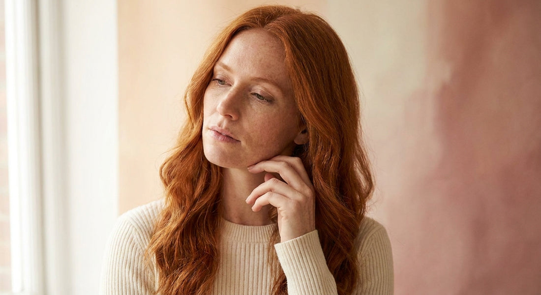A fair-skinned redhead woman with copper hair and visible freckles touches her face thoughtfully against a warm cream background, evoking skin sensitivity and self-awareness.