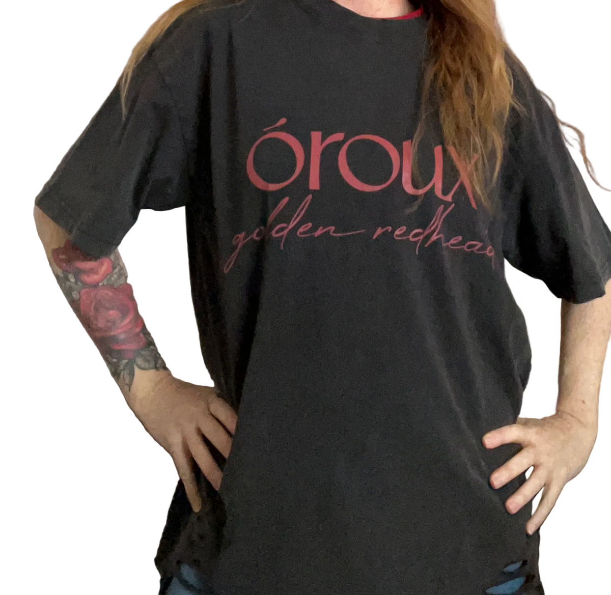 A person wearing an oversized black graphic T-shirt with pink text reading 'oroux golden redhead' across the front, standing indoors with hands on hips.
