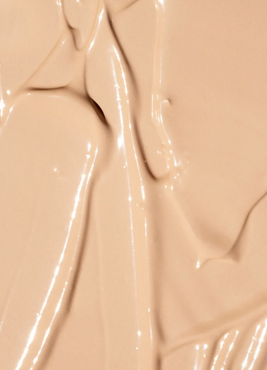 A close-up of a smooth, glossy beige cosmetic cream or foundation swatch with soft ridges and reflective highlights.