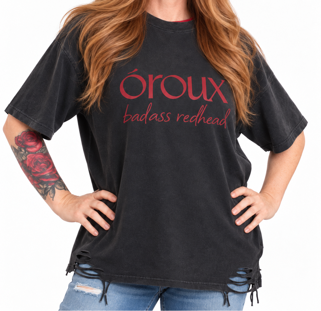 A woman with long red hair wearing a faded black graphic t-shirt with 'oroux badass redhead' printed in red on the front, paired with blue jeans against a white background.