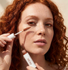 A close-up of a woman with curly red hair applying a redhead mascara cosmetic product from a tube while holding the applicator wand. The brand name &