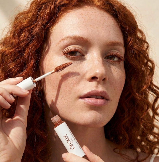 A close-up of a woman with curly red hair applying a redhead mascara cosmetic product from a tube while holding the applicator wand. The brand name 'OROUX' is visible on the tube.