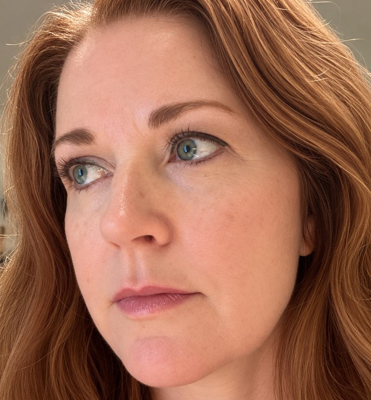 Close-up portrait of a woman with red hair and blue eyes, looking off to the side, showing natural skin texture and makeup. Wearing Long and Roux Mascara for redheads by Óroux skin and Roux eyebrow pencil for redheads and auburn