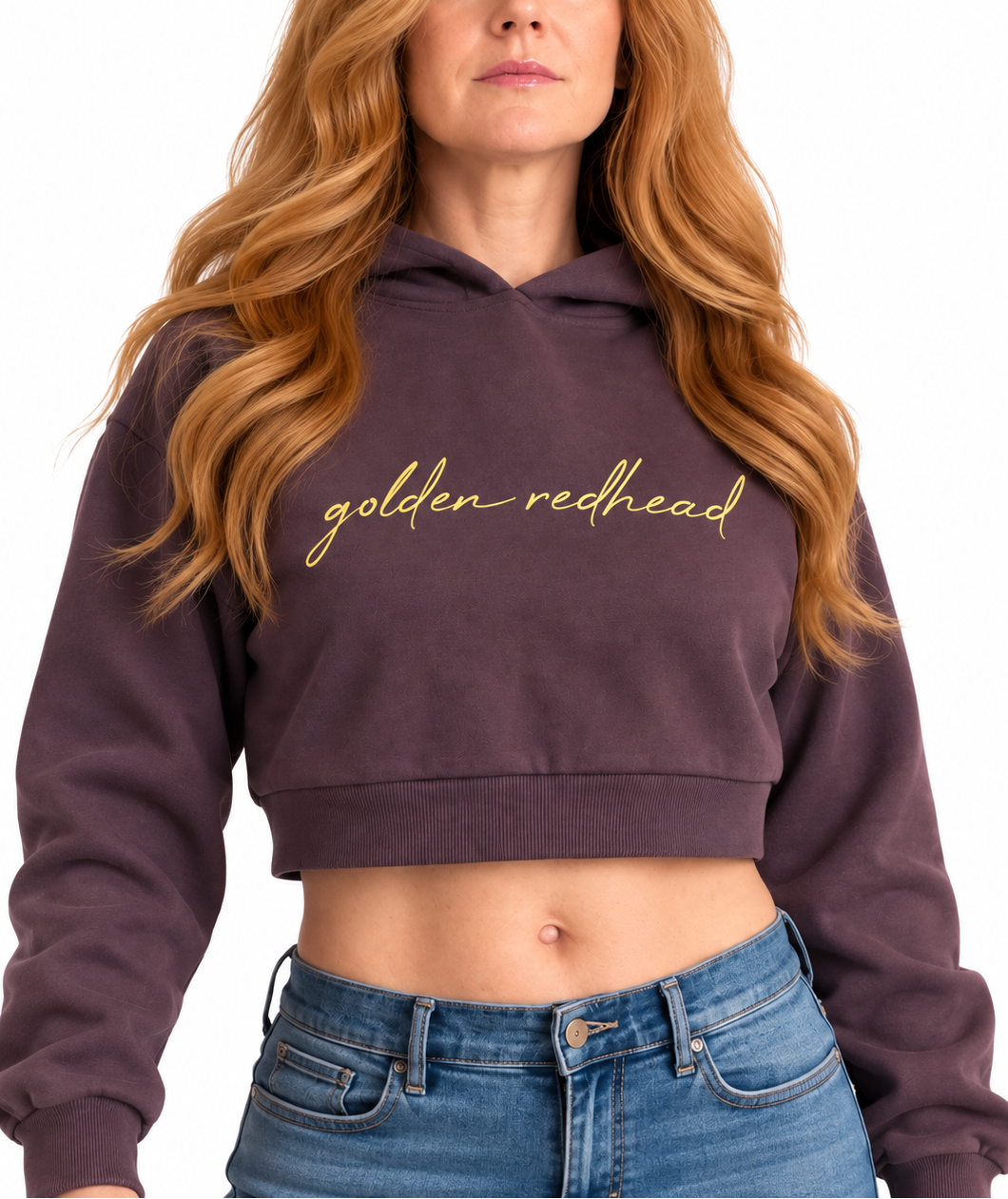 A woman with long wavy auburn hair wearing a dark plum purple cropped hoodie with 'golden-redhead' embroidered in gold script across the chest, paired with blue jeans, against a plain white background.