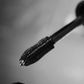 Close-up black and white photo of a mascara wand brush tip above an open tube, with no person or real environment visible.
