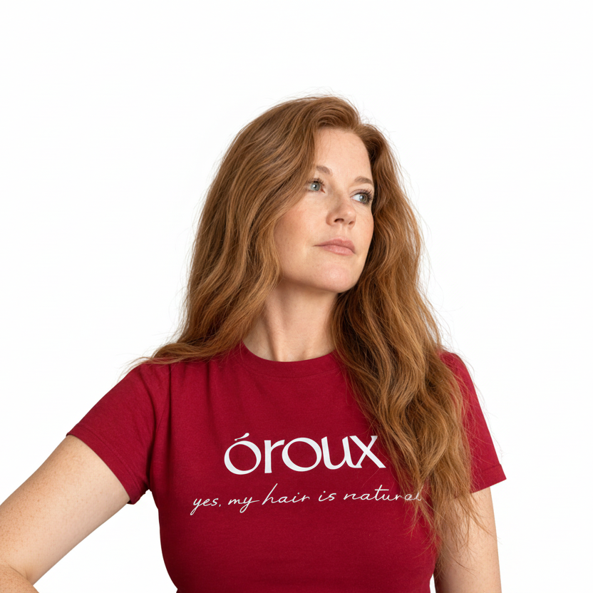 A woman with long wavy light red hair wearing a red t-shirt with white text that reads 'OROUX' and 'yes my hair is natural' in cursive, photographed indoors against a plain light wall.