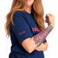 A split-screen lifestyle image of a woman with long red hair modeling a navy blue t-shirt. The left side shows her from the waist up with her hand on her hip, and the right side shows a closer cropped view of her flexing her arm.