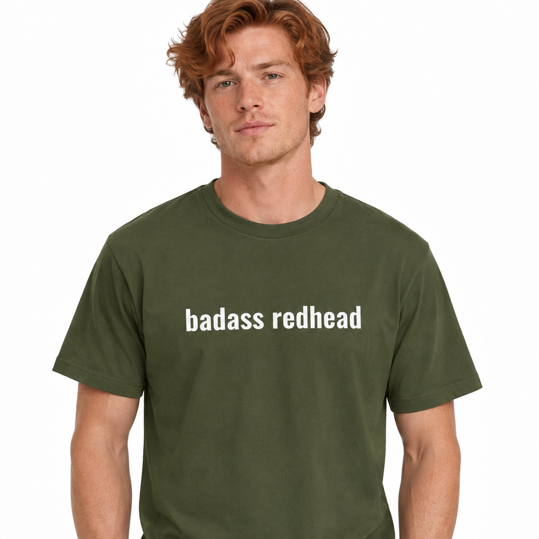A man wearing an olive green crewneck t-shirt with the text 'badass redhead' printed in white across the chest, paired with black pants, against a plain white background.