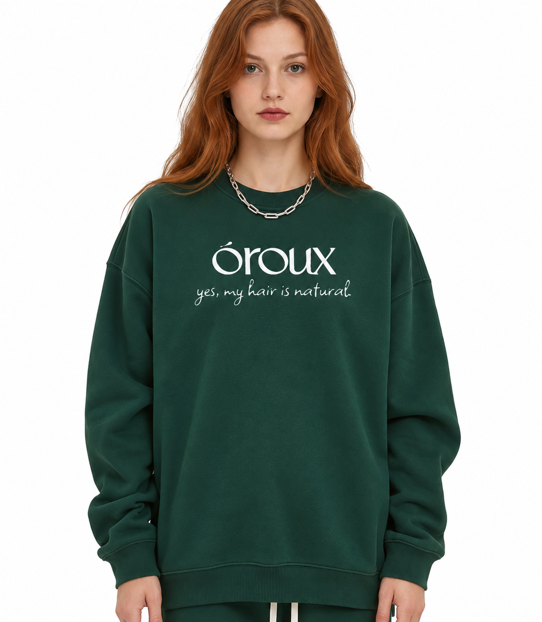 A woman with red hair wearing a dark green crewneck sweatshirt with white text that reads 'oroux yes, my hair is natural.' and matching green sweatpants, photographed against a plain white background.
