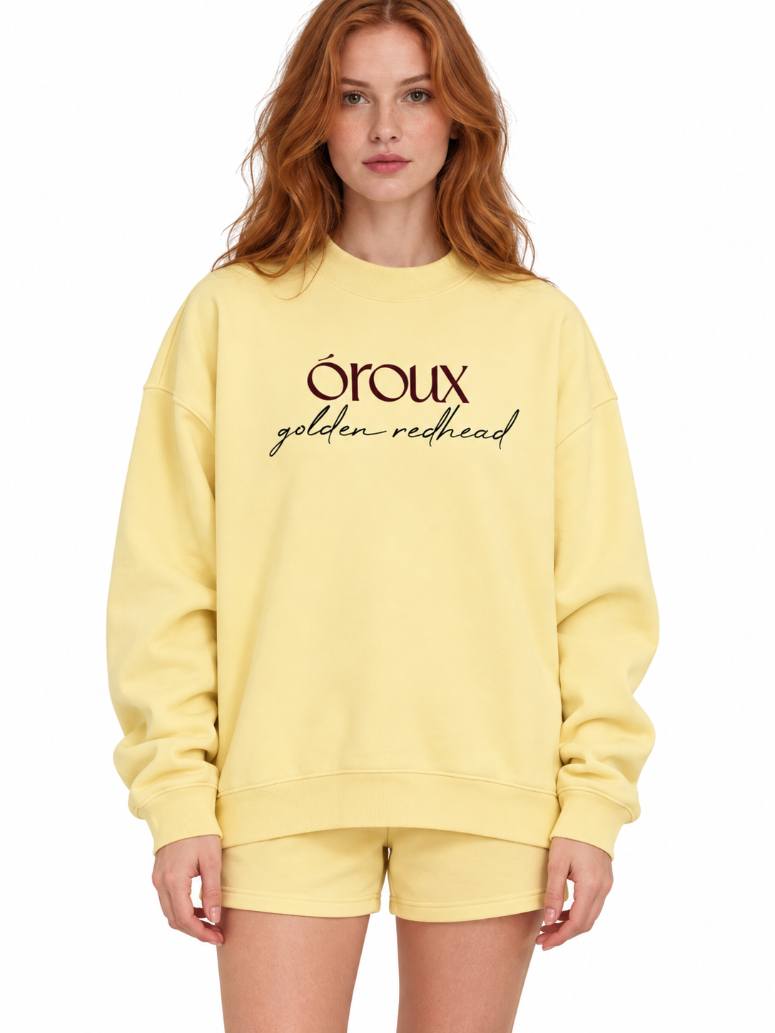 A woman with red hair wearing a pale yellow sweatshirt and matching shorts. The sweatshirt features the text 'oroux golden redhead' printed in dark brown across the chest.