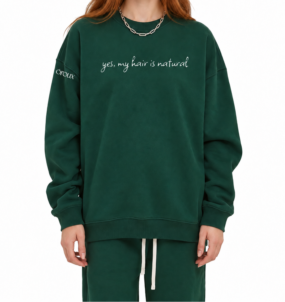A model with red hair wearing a dark green crewneck sweatshirt with the phrase 'yes, my hair is natural' printed in white script across the chest, paired with matching green sweatpants against a plain white background.
