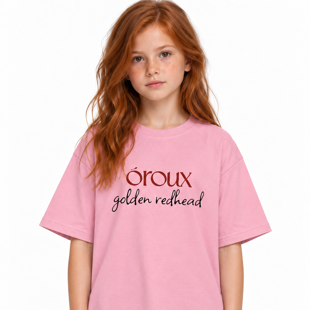 A young girl with red hair wearing a pink t-shirt with 'oroux golden redhead' printed on the chest, paired with black pants, against a plain white background.